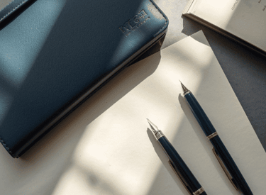 A blue leather notebook and two luxury fountain pens resting on paper in soft natural sunlight.