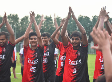 Reeds football club players in red and black jerseys clapping on a grass soccer field.
