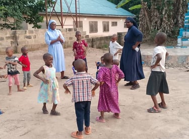 Two Catholic nuns playing outdoors with a group of young African children in a village courtyard.