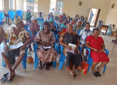 A community meeting of African women sitting in blue plastic chairs at an indoor workshop.