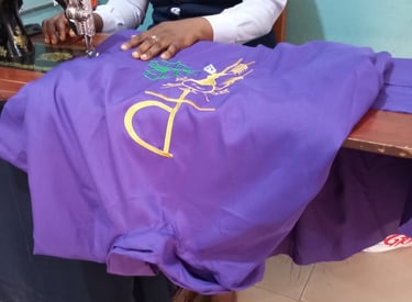 A tailor using a sewing machine to embroider a gold religious symbol onto purple liturgical fabric.