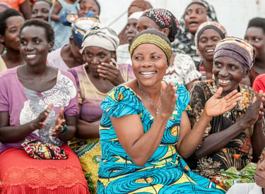 A group of smiling African women wearing colorful traditional clothing while clapping at a community gathering.