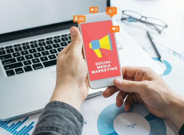 A person holding a smartphone displaying a social media marketing graphic next to a laptop and data charts.