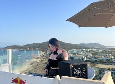 A female DJ performs at a rooftop beach club overlooking the coast with a Pioneer speaker and Red Bull booth.