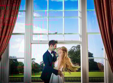A happy bride and groom embrace in front of a large window overlooking a scenic garden estate.