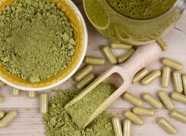 Organic green kratom powder in a bowl and wooden scoop with herbal capsules on a table.