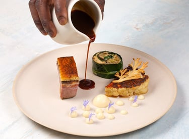 Chef pouring dark jus over a gourmet plate of roasted pork belly, stuffed cabbage, and cauliflower steak.