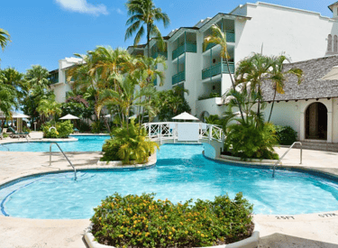 Luxury tropical resort pool with a white footbridge, palm trees, and bright blue water under a clear sky.