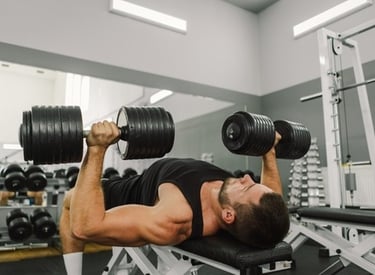 A muscular man performing a chest press with heavy dumbbells on a flat bench in a modern gym.