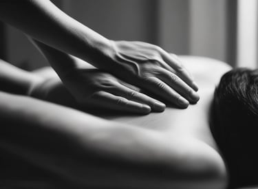 A person is giving a massage in a dimly lit room. The focus is on the hands applying pressure on the back of the individual lying down. The atmosphere appears calm and therapeutic.