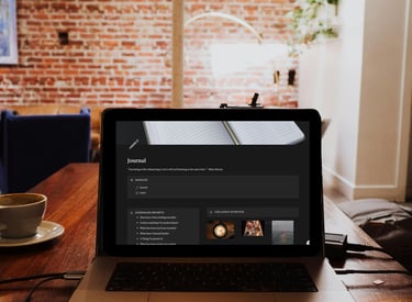 A laptop on a wooden desk displays a digital journal app next to a cup of coffee in a brick-walled office.