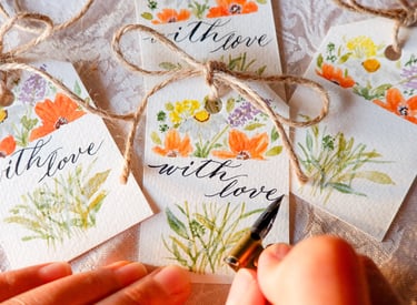 a calligrapher writing the words “with love” for a gift tag with handmade watercolor florals