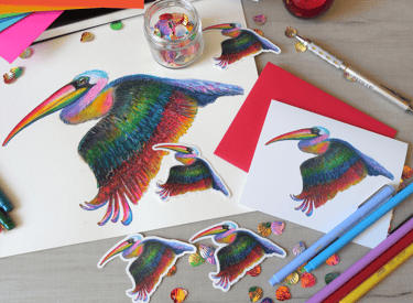Hand-drawn rainbow pelican illustration with stickers and colorful envelope on a desk.