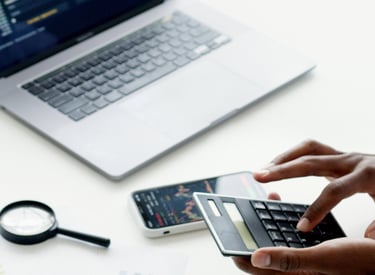 Pracownik dokonujący analiz finansowych na kalkulatorze, obok laptopa z wykresami.