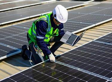 Engineer in safety gear using a laptop to inspect solar panel installation on a rooftop.