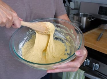 A person using a wooden spoon to mix thick yellow bread dough in a glass bowl.