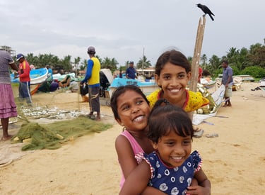 Three smiling Sri Lankan girls playing on a sandy beach near fishing boats and nets in a coastal village.