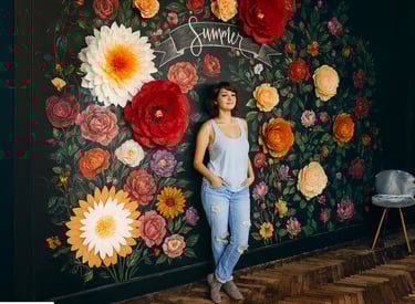 In the image, a young woman is standing against a dark wall decorated with a colorful floral mural. The mural features many l