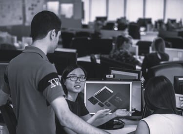 three colleagues, two sat by a computer with another stood with them discussing options for a projec