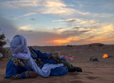 A man in traditional dress lies in the desert watching the sunset