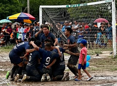 Muddy soccer players in blue jerseys celebrate a goal on a dirt field during a community football match.