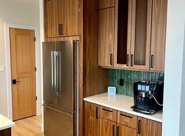 Modern kitchen with wood cabinets, stainless steel refrigerator, and a coffee maker on white counters.