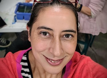 A smiling woman with brown eyes and hair wearing a bright pink cardigan in an art studio.