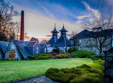 Historic Scottish whisky distillery with pagoda roofs and tall chimney at sunset.
