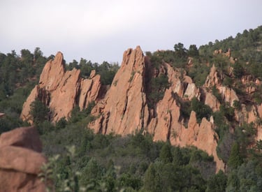 Towering red rock sandstone formations rise above a lush green forest in a scenic mountain landscape.