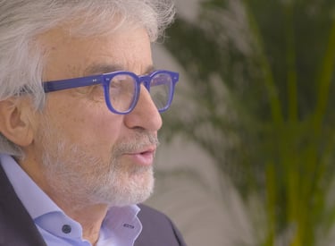A senior professional man with grey hair and blue glasses speaking in a modern office.
