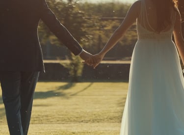 A bride and groom holding hands while walking through a sunny garden during an outdoor wedding ceremony.