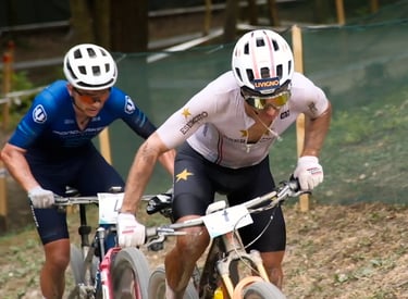 Two professional cross-country mountain bikers racing uphill on a dirt trail wearing helmets and cycling kits.
