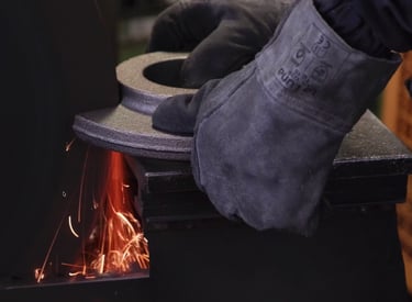 Gloved worker grinding a metal industrial part on a wheel with sparks flying.