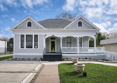 Restored light gray Victorian cottage with a white porch railing and yellow front door.