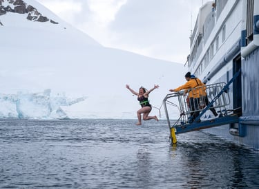 A tourist does a polar plunge from a cruise ship into icy Antarctic waters near snowy mountains.