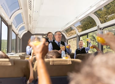 Passengers and staff toasting with orange juice in a luxury dome train car with panoramic windows.