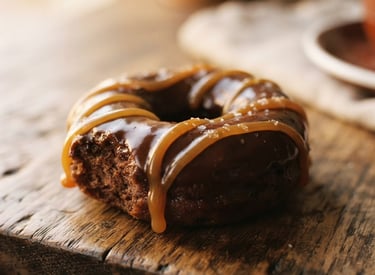 Warm editorial food photography, close-up macro of a freshly glazed chocolate donut with golden caramel drizzle, resting on a rustic wooden table with a soft golden window light glow, shallow depth of field, 35mm.