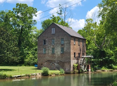 A historic brick watermill with a large wooden wheel sits beside a calm river in a lush green forest.
