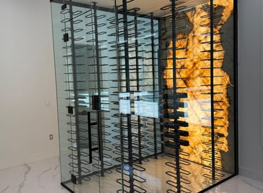 Modern glass wine cellar with black metal racks and a striking backlit onyx or marble accent wall for elegant wine storage.