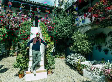 A man posing on stairs in a flower-filled traditional Cordoba courtyard with blue pots and blooming plants.