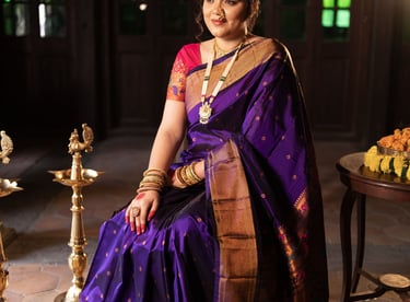 Indian woman in a purple Paithani silk saree with gold jewelry sitting in a traditional setting.