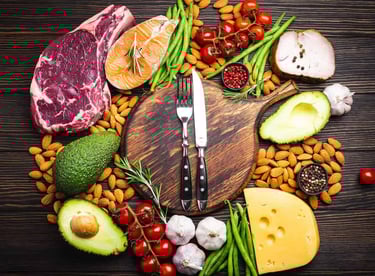  Alimentos variados como carne, peixe, queijo, abacate e vegetais em torno de tábua de madeira.