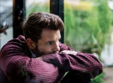 Un homme mal dans sa peau est assis au sol bras croisés, et observe le monde extérieur par la fenêtre.
