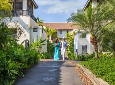 A couple walking down a tropical resort pathway lined with lush greenery, ferns, and palm trees