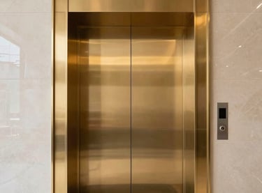 A low-angle, professional architectural photograph of a sleek, modern elevator entrance in a South Asian corporate headquarters. The doors are finished in polished gold, reflecting a minimalist lobby with black marble floors and soft, premium lighting. The composition is centered and symmetrical, projecting corporate reliability and precision.
