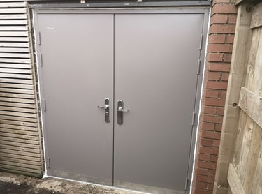 Durable grey steel double security doors installed between a brick wall and wooden fencing.