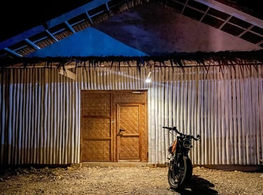 A vintage motorcycle parked at night in front of a rustic bamboo house with woven doors.