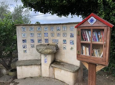 A stone drinking fountain with decorative tiles next to a wooden Little Free Library box filled with books.