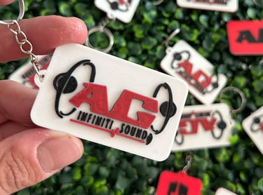 Custom 3D printed AG Infiniti Sound logo keychain held against a green hedge background.