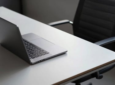 A minimalist workspace featuring a laptop on a light grey desk. A dark charcoal chair is partially visible. The scene is organized and professional, reflecting clear business operations and workflows.
